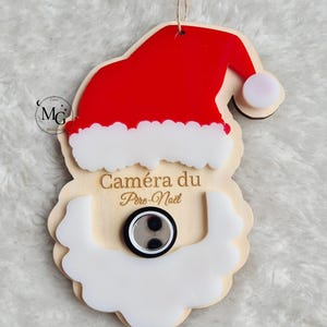 Peut inclure: Ornement de Noël en bois représentant le visage du Père Noël, avec un bonnet rouge, une barbe blanche et le texte "Caméra du Père Noël". L'ornement comprend un petit détail de caméra rond, noir et argenté.