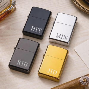 Può includere: Quattro accendini personalizzati in nero, argento e oro. Ogni accendino ha un'iscrizione diversa di tre lettere: "HIT", "MIN", "KIB" e "JID". Gli accendini sono esposti su una superficie di legno chiaro, con una bottiglia di whisky e un sigaro.