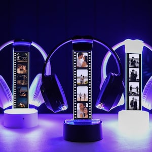 May include: Three illuminated headphone displays with personalized photo strips. The headphones are black and white, with a strip of photos in the center. The bases are white and black, with a purple glow. The background is dark.