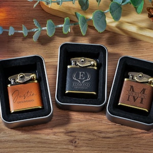 Può includere: Tre accendini personalizzati in custodie metalliche. Gli accendini hanno un aspetto simile alla pelle con dettagli in metallo color oro. Ogni accendino è inciso con un nome o iniziali. Gli accendini sono esposti su una superficie di legno.