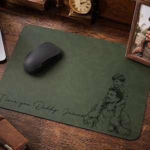 May include: A rectangular olive green desk mat featuring a black line art illustration of a person carrying a child on their shoulders. The text "I love you Daddy, James" is printed in a cursive script on the left side of the mat.