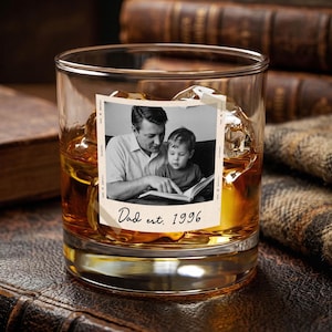 May include: A clear tumbler filled with amber liquid and ice cubes. A black and white photograph is attached to the glass, showing a father and child reading a book. The text "Dad est. 1996" is below the photo.