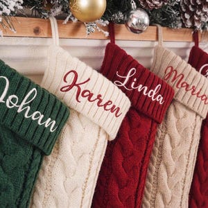 Op de afbeelding: Vijf kerstsokken van kabelbreisel hangen op een rij, afwisselend in de kleuren donkergroen, crème en rood. Elke sok heeft een geribbelde boord die is geborduurd met de namen Johan, Karen, Linda, Maria en Crystal in een cursief lettertype.