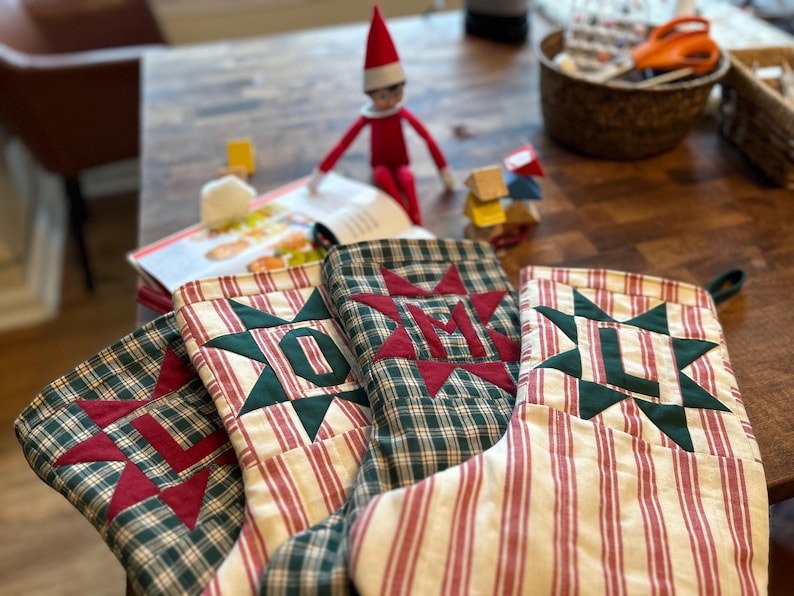 May include: Three fabric Christmas stockings featuring a red and green plaid pattern with a cream and red striped cuff. Each stocking displays a large, dark green star appliqu&eacute; centered with a red fabric letter: L, O, and M respectively.