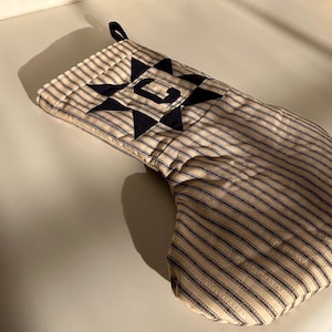 May include: A quilted Christmas stocking featuring a beige and navy blue ticking stripe pattern. The cuff is decorated with a navy blue fabric star appliqu&eacute; centered with the capital letter C in a matching navy blue fabric.