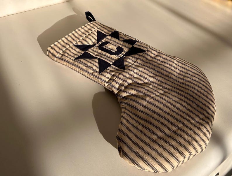 May include: A quilted Christmas stocking featuring a beige and navy blue ticking stripe pattern. The cuff is decorated with a navy blue fabric star appliqu&eacute; centered with the capital letter C in a matching navy blue fabric.