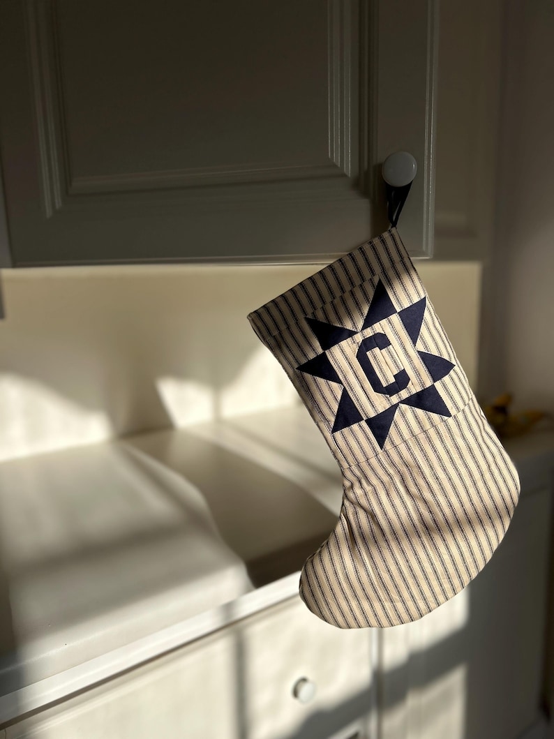 May include: A Christmas stocking made of beige and navy blue striped fabric. The stocking features a navy blue star-shaped appliqu&eacute; on the cuff with the letter C centered in the middle of the star. The stocking is hanging from a cabinet knob.