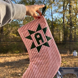 May include: A Christmas stocking featuring a red and white vertical striped fabric pattern. The stocking is decorated with a dark green fabric star  centered on the front, which contains a dark green letter A in the middle.