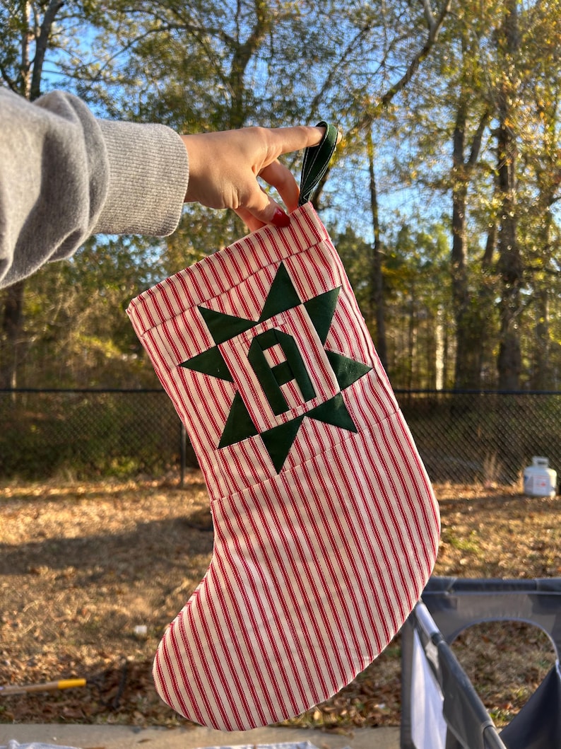 May include: A Christmas stocking featuring a red and white vertical striped fabric pattern. The stocking is decorated with a dark green fabric star  centered on the front, which contains a dark green letter A in the middle.