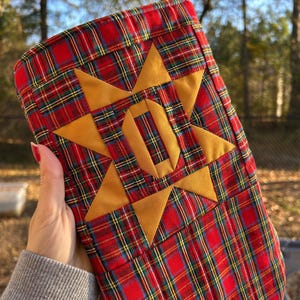 May include: A Christmas stocking made of red, green, and yellow plaid fabric featuring a large, centered, eight-pointed mustard yellow star. The stocking includes a matching mustard yellow fabric hanging loop at the top edge.