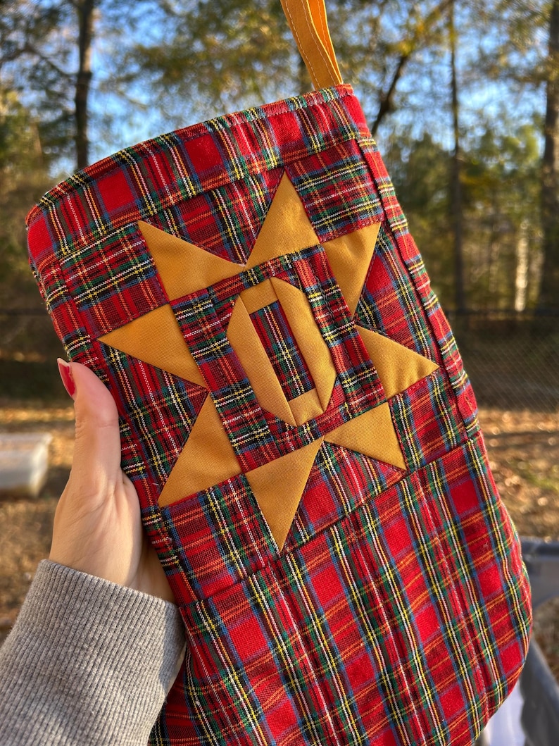 May include: A Christmas stocking made of red, green, and yellow plaid fabric featuring a large, centered, eight-pointed mustard yellow star. The stocking includes a matching mustard yellow fabric hanging loop at the top edge.