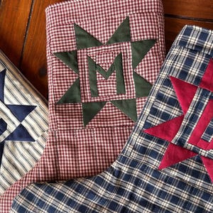 May include: Three fabric Christmas stockings featuring star-shaped appliques with centered letters. One stocking is cream with navy stripes and a navy C, one is red and white gingham with a green M, and one is navy plaid with a red D.