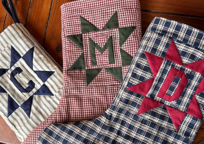 May include: Three fabric Christmas stockings featuring star-shaped appliques with centered letters. One stocking is cream with navy stripes and a navy C, one is red and white gingham with a green M, and one is navy plaid with a red D.