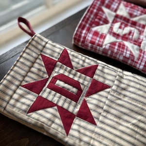 May include: A quilted fabric stocking featuring a cream and black ticking stripe pattern with a central dark red star appliqu&eacute; containing the letter D. A maroon fabric loop is attached to the top corner for hanging purposes.