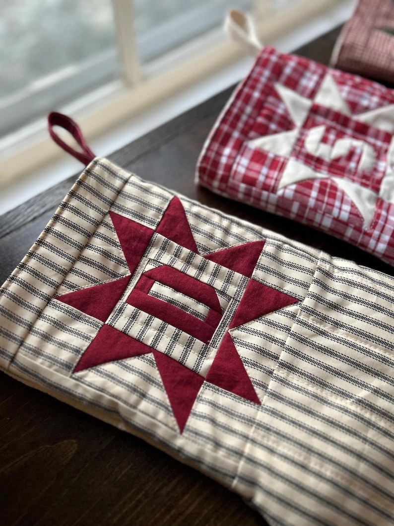 May include: A quilted fabric stocking featuring a cream and black ticking stripe pattern with a central dark red star appliqu&eacute; containing the letter D. A maroon fabric loop is attached to the top corner for hanging purposes.