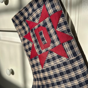 May include: A Christmas stocking made of navy blue and beige plaid fabric. The stocking features a large, red, eight-pointed star appliqu&eacute; centered on the front, with a bold, red, block-style letter D stitched directly into the star's center.