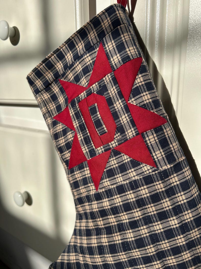 May include: A Christmas stocking made of navy blue and beige plaid fabric. The stocking features a large, red, eight-pointed star appliqu&eacute; centered on the front, with a bold, red, block-style letter D stitched directly into the star's center.