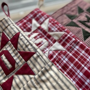May include: Three fabric stockings featuring quilted star patterns. The first is cream with black stripes and a dark red star. The second is a red and white plaid with a cream star. The third is a red and white gingham with a dark green star.