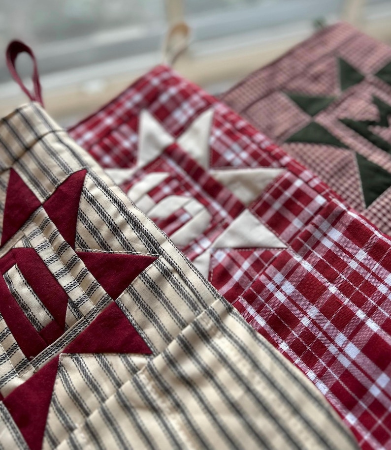 May include: Three fabric stockings featuring quilted star patterns. The first is cream with black stripes and a dark red star. The second is a red and white plaid with a cream star. The third is a red and white gingham with a dark green star.