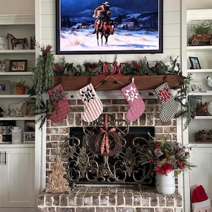 May include: A brick fireplace features four hanging Christmas stockings with letters H, J, and M, topped by a wooden mantel with greenery and a red JOY sign. Above, a television displays a cowboy on a horse in a snowy landscape.