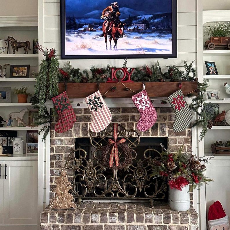 May include: A brick fireplace features four hanging Christmas stockings with letters H, J, and M, topped by a wooden mantel with greenery and a red JOY sign. Above, a television displays a cowboy on a horse in a snowy landscape.