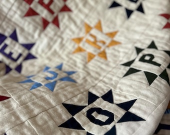May include: Close-up of a white quilt with a star pattern. Each star contains a letter, with colors including red, blue, yellow, and navy. The quilt has a quilted texture and a soft, inviting appearance.