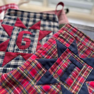 May include: Two quilted fabric Christmas stockings featuring plaid patterns and star appliques. One stocking is navy and cream plaid with a red star and the letter G, while the other is red, green, and yellow plaid with a navy blue star.