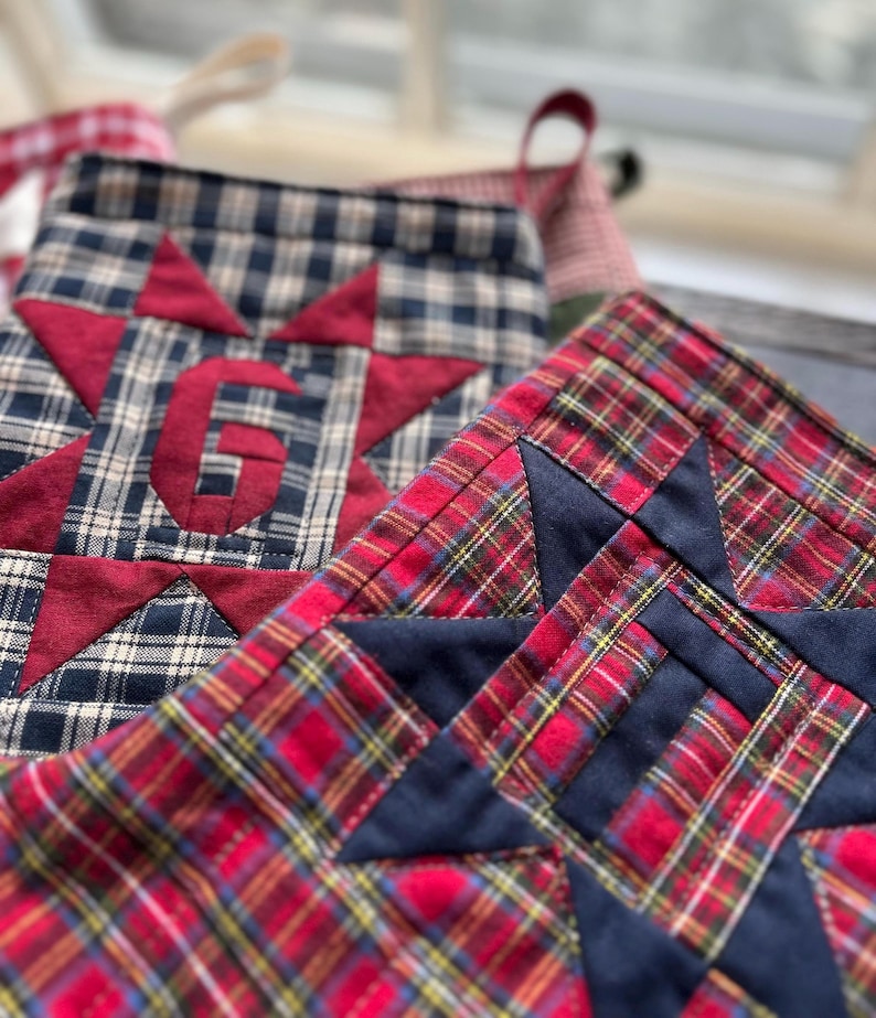 May include: Two quilted fabric Christmas stockings featuring plaid patterns and star appliques. One stocking is navy and cream plaid with a red star and the letter G, while the other is red, green, and yellow plaid with a navy blue star.