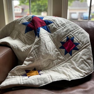 May include: Cream-colored quilt with star patterns in red, blue, and yellow. The quilt is folded over a brown leather couch, with a window in the background. The stars are of varying sizes and are evenly spaced across the quilt.
