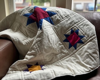 May include: Cream-colored quilt with star patterns in red, blue, and yellow. The quilt is folded over a brown leather couch, with a window in the background. The stars are of varying sizes and are evenly spaced across the quilt.
