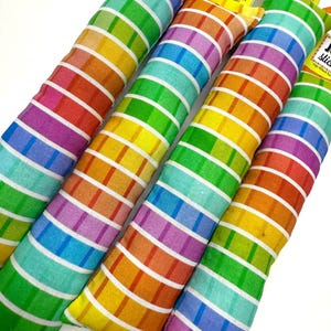 Op de afbeelding: Vier cilindrische speeltjes met regenboogstrepen. Elk speeltje heeft een witte achtergrond met rode, oranje, gele, groene, blauwe en paarse strepen. Kleine gele labels dragen de tekst "JOY stick" in zwarte letters.