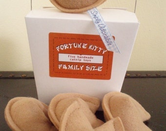 May include: A white box with the text "Fortune Kitty" and "Family Size" on it. The box contains five handmade catnip toys shaped like fortune cookies. The toys are beige and have a blue ribbon with the text "Simply Adorable" on it.