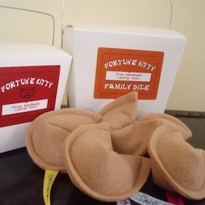 May include: Two white boxes with red labels that say "Fortune Kitty" and contain catnip toys. The smaller box says "three handmade catnip toys" and the larger box says "five handmade catnip toys FAMILY SIZE". There are five brown felt catnip toys shaped like fortune cookies.