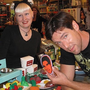 May include: Two people are standing in a store. The person on the left is wearing a black shirt and has short blonde hair. The person on the right is wearing a black t-shirt with a cat graphic and is holding a small, round, colorful sticker with a person's face on it. The sticker is on a table with other items, including a blue metal stand with two bowls, a white box, and a red and green toy.