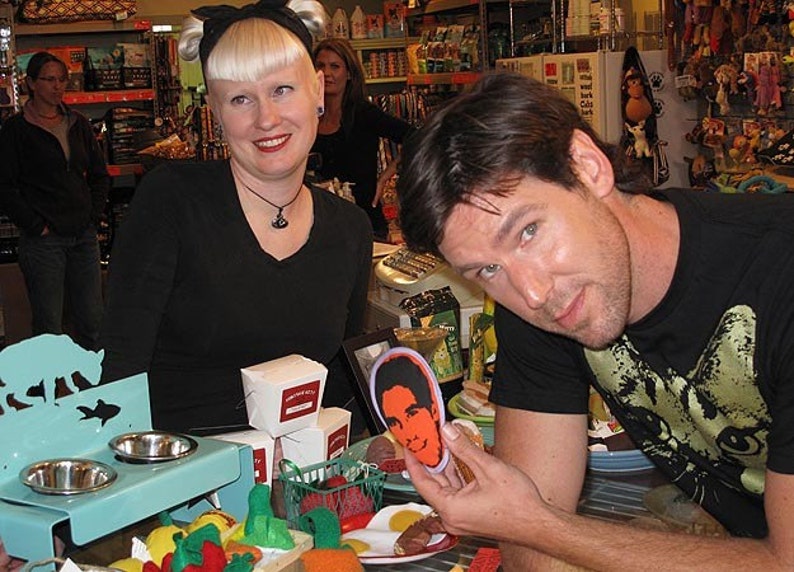 May include: Two people are standing in a store. The person on the left is wearing a black shirt and has short blonde hair. The person on the right is wearing a black t-shirt with a cat graphic and is holding a small, round, colorful sticker with a person's face on it. The sticker is on a table with other items, including a blue metal stand with two bowls, a white box, and a red and green toy.