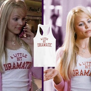 May include: A white tank top with the text "A Little Bit Dramatic" in red lettering. The tank top is worn by a person with blonde hair and a pink cardigan.