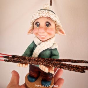 May include: A charming elf figurine, dressed in a green jacket, white scarf, and a cream-coloured hat. The elf is holding several brown sticks with red tips. The figurine stands on a wooden base. The text "Pomelo Porcelana" is visible on the base.