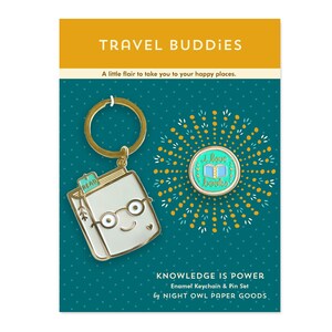 May include: A gold enamel keychain and pin set featuring a book with glasses and the text "Read". The pin features a turquoise background with the text "I love books" and a pair of open books.