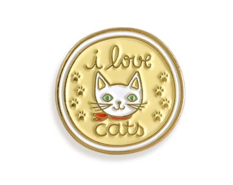 Puede incluir: Un pin de esmalte dorado con un gato blanco que lleva una pajarita roja. El pin tiene un fondo amarillo con el texto "i love cats" y huellas de patas alrededor del gato.