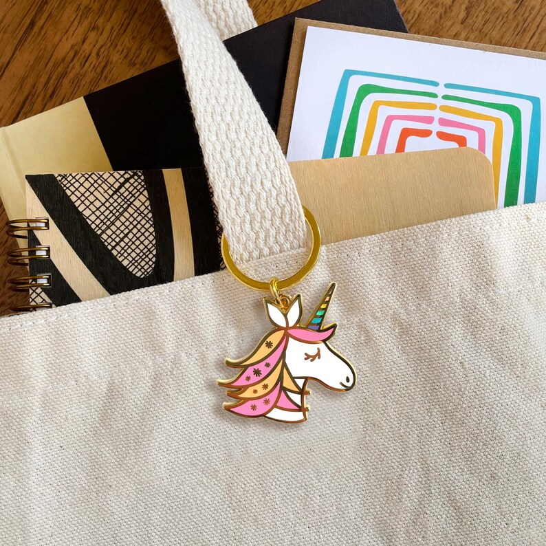 May include: A gold-tone keychain with a colorful unicorn charm. The unicorn has a pink, yellow, and blue mane and tail with white flowers. The unicorn's horn is rainbow colored.