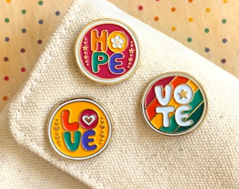 May include: Three colorful enamel pins with gold borders. One pin reads "HOPE" with a flower, another says "LOVE" with a heart, and the third says "VOTE" with a star. The pins are on a beige fabric.