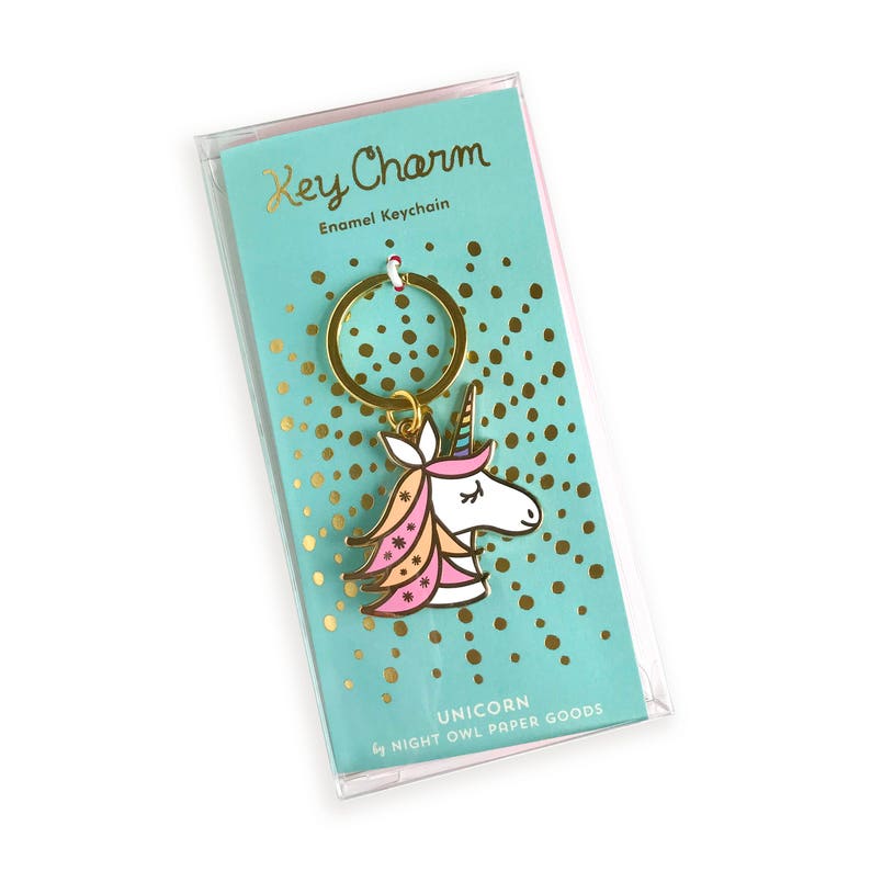 May include: A gold key chain with a pink, white, and yellow unicorn charm. The charm has a gold horn and a gold ring. The packaging says "Key Charm Enamel Keychain Unicorn by Night Owl Paper Goods".
