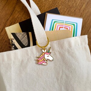 May include: A white canvas tote bag with a gold unicorn keychain. The unicorn has pink, yellow, and blue details. The bag is filled with notebooks and cards.