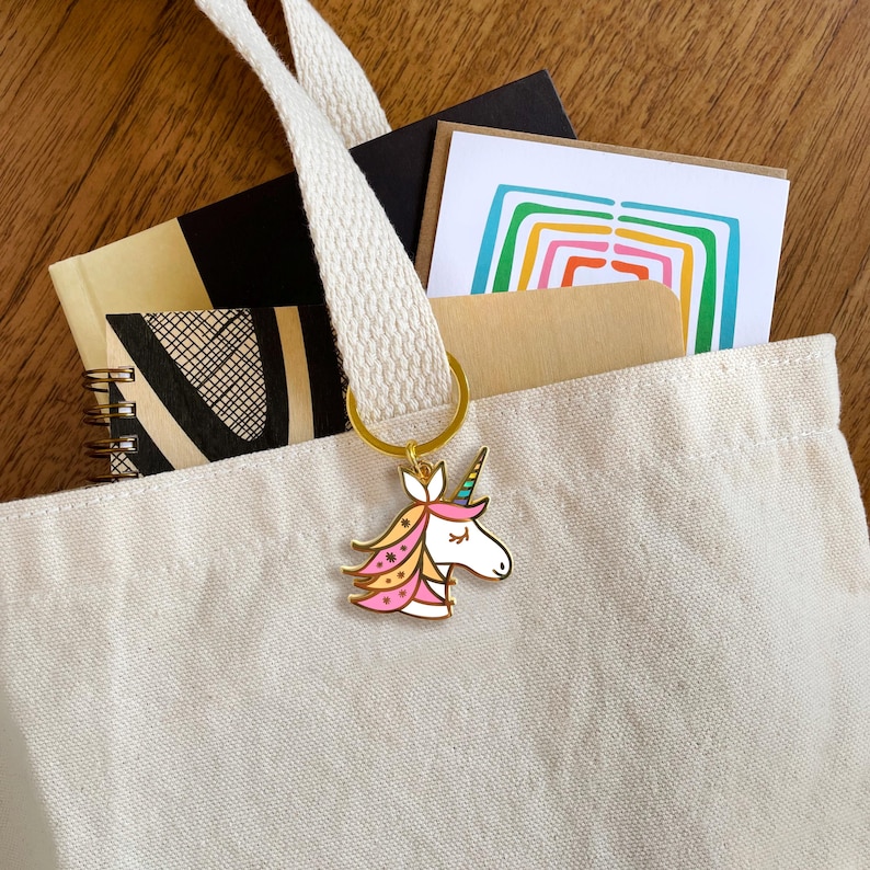 May include: A white canvas tote bag with a gold unicorn keychain. The unicorn has pink, yellow, and blue details. The bag is filled with notebooks and cards.