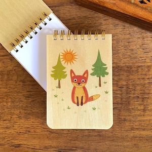 May include: A small wooden notepad with a spiral binding features a cartoon fox in a forest setting. The fox is orange and red with a white belly and a black nose. The forest is green with a yellow sun in the background.