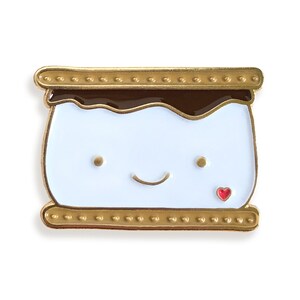 May include: Gold-tone enamel pin shaped like a smiling s'more with a red heart. The s'more has a white marshmallow, brown chocolate, and a golden graham cracker.