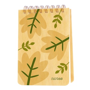 May include: A spiral-bound notepad with a light wood-grain cover. The cover features a repeating pattern of large, light yellow leaves with green veins and smaller green leaf sprigs. The word "notes" is printed in a small, dark font at the bottom.