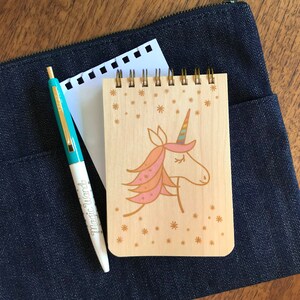 May include: A wooden notepad with a unicorn illustration, a teal and gold pen with the words "Magic Wand", and a piece of white paper, all resting on a dark blue denim pouch. The notepad has a spiral binding and a light wood finish.