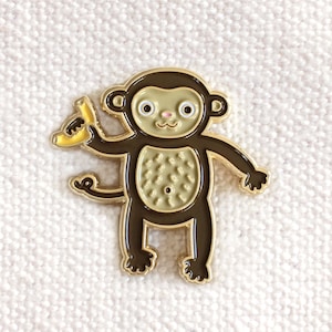 May include: A gold-colored enamel pin featuring a cartoon monkey holding a banana. The monkey has brown fur, a light brown belly, and a pink nose. The banana is yellow with brown spots.