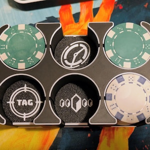 May include: A black plastic chip holder with six circular compartments, each holding a game chip. The chips are teal, white, and black, with various designs including a target, a compass, and the word "TAG".